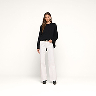 A woman stands against a plain white background wearing the Loden Tunic Pullover in a relaxed fit, paired with white wide-leg pants, black boots, and hoop earrings. Her hair is down, and one hand is in her pocket.