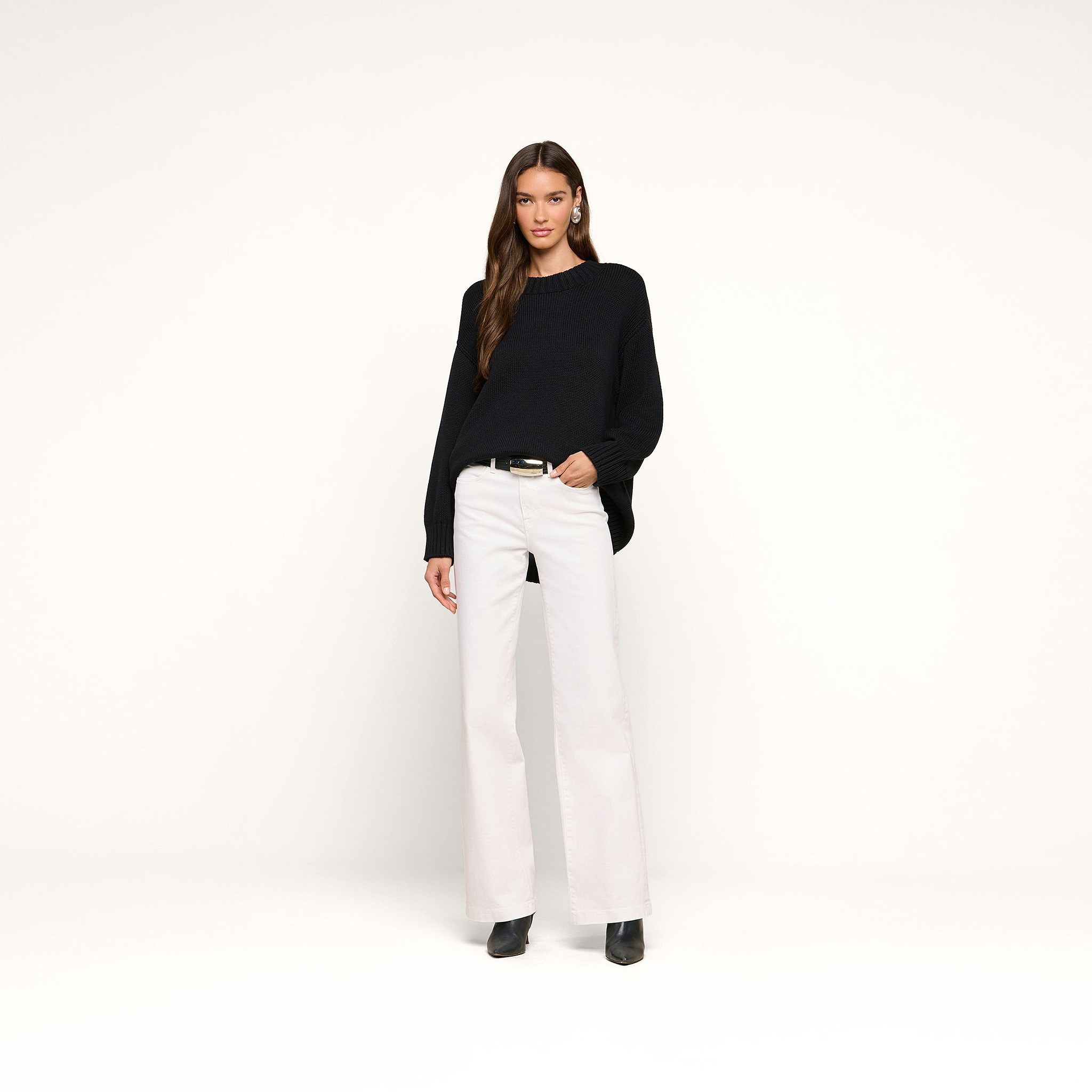 A woman stands against a plain white background wearing the Loden Tunic Pullover in a relaxed fit, paired with white wide-leg pants, black boots, and hoop earrings. Her hair is down, and one hand is in her pocket.