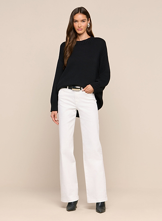 A woman stands against a beige background, wearing the Loden Pullover—an everyday black sweater in a relaxed fit styled as a loose pullover—paired with white wide-leg pants, black ankle boots, and a black belt. She has long brown hair and looks at the camera with a slight smile.