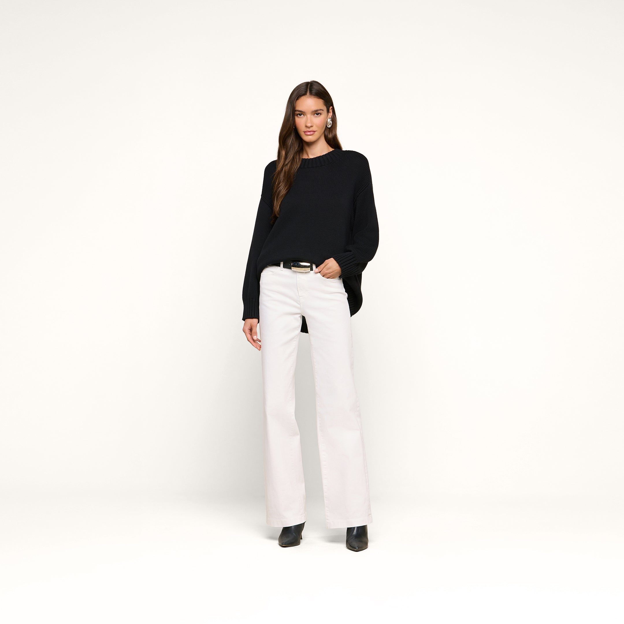 A woman stands against a plain white background wearing the Loden Tunic Pullover in a relaxed fit, paired with white wide-leg pants, black boots, and hoop earrings. Her hair is down, and one hand is in her pocket.