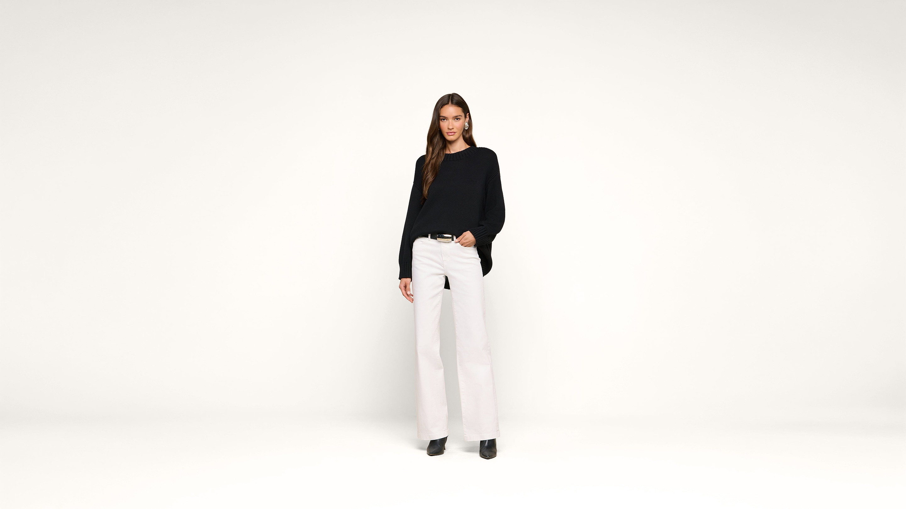 A woman with long brown hair wearing a black sweater, white wide-leg pants, and black shoes stands against a plain white background, looking at the camera and smiling slightly.