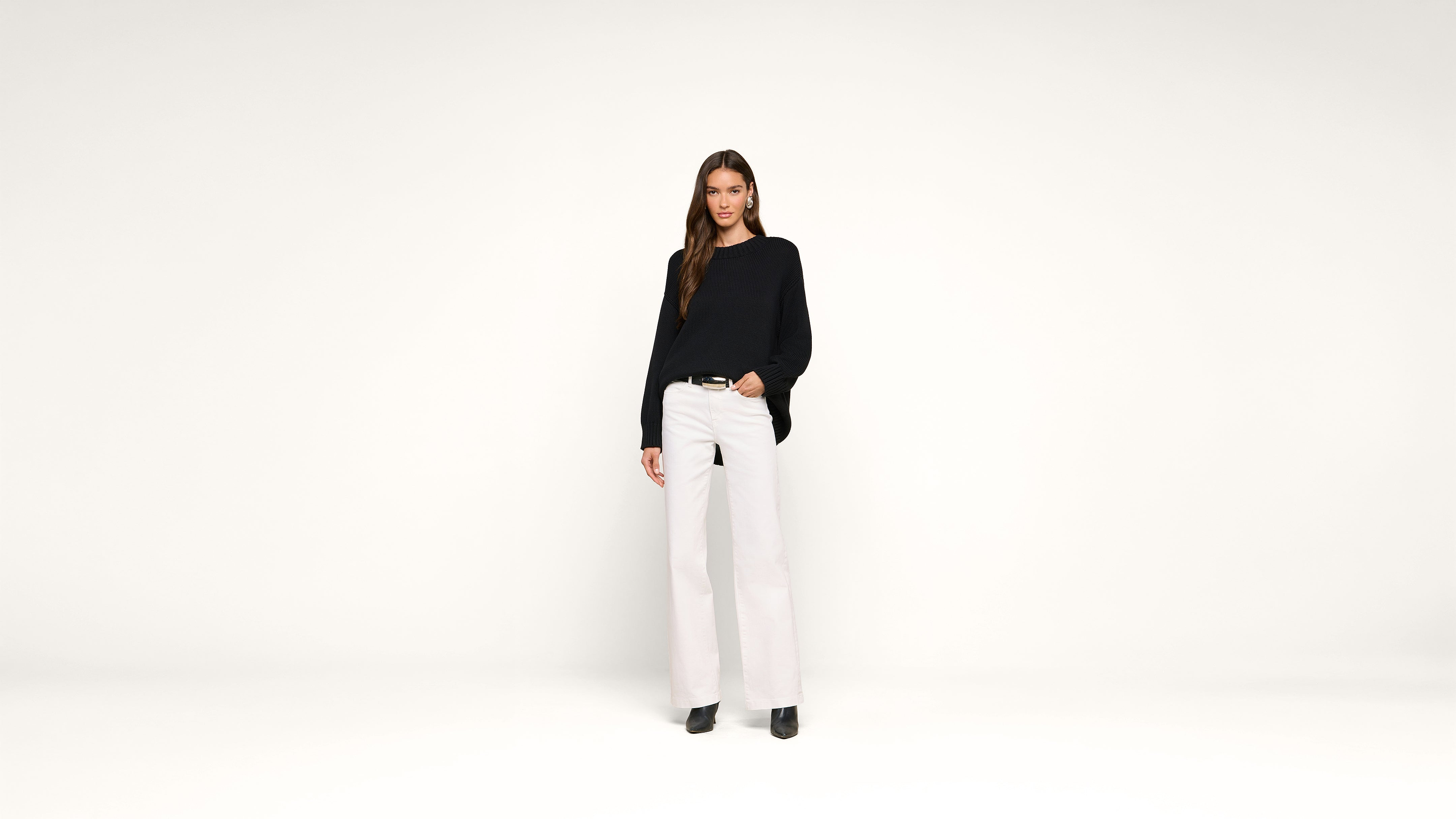 A woman with long brown hair wearing a black sweater, white wide-leg pants, and black shoes stands against a plain white background, looking at the camera and smiling slightly.