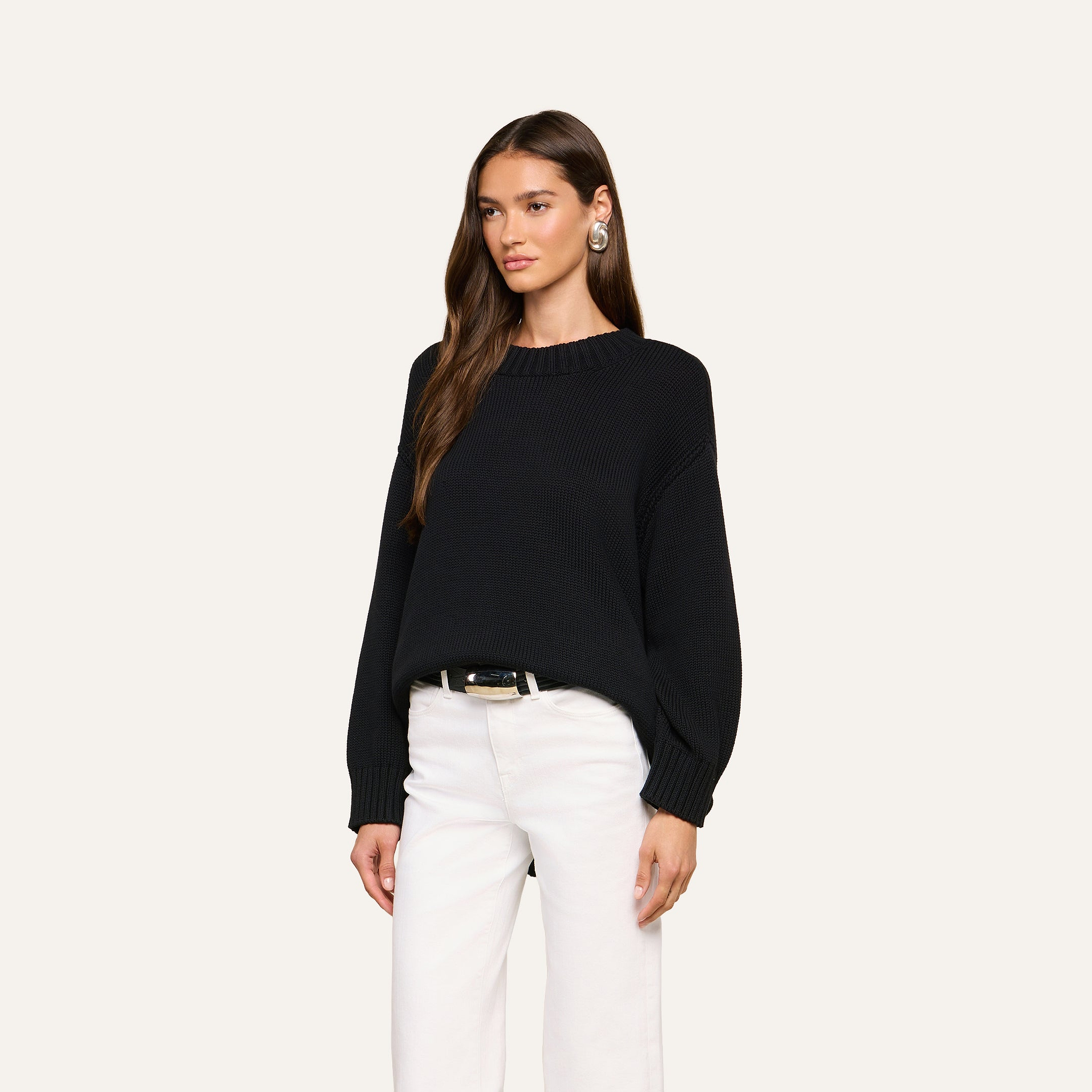 A woman with long brown hair wears the Loden Tunic Pullover, a relaxed-fit black everyday sweater, paired with white pants and large earrings. She stands against a plain, light background, looking slightly to the side with a neutral expression.