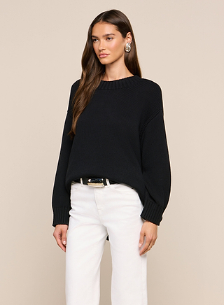A woman with long, straight brown hair and a neutral expression stands against a plain background, wearing the Loden Pullover—a relaxed fit black sweater with long sleeves—paired with white pants and large round earrings. The Loden Pullover offers comfort and style for any occasion.