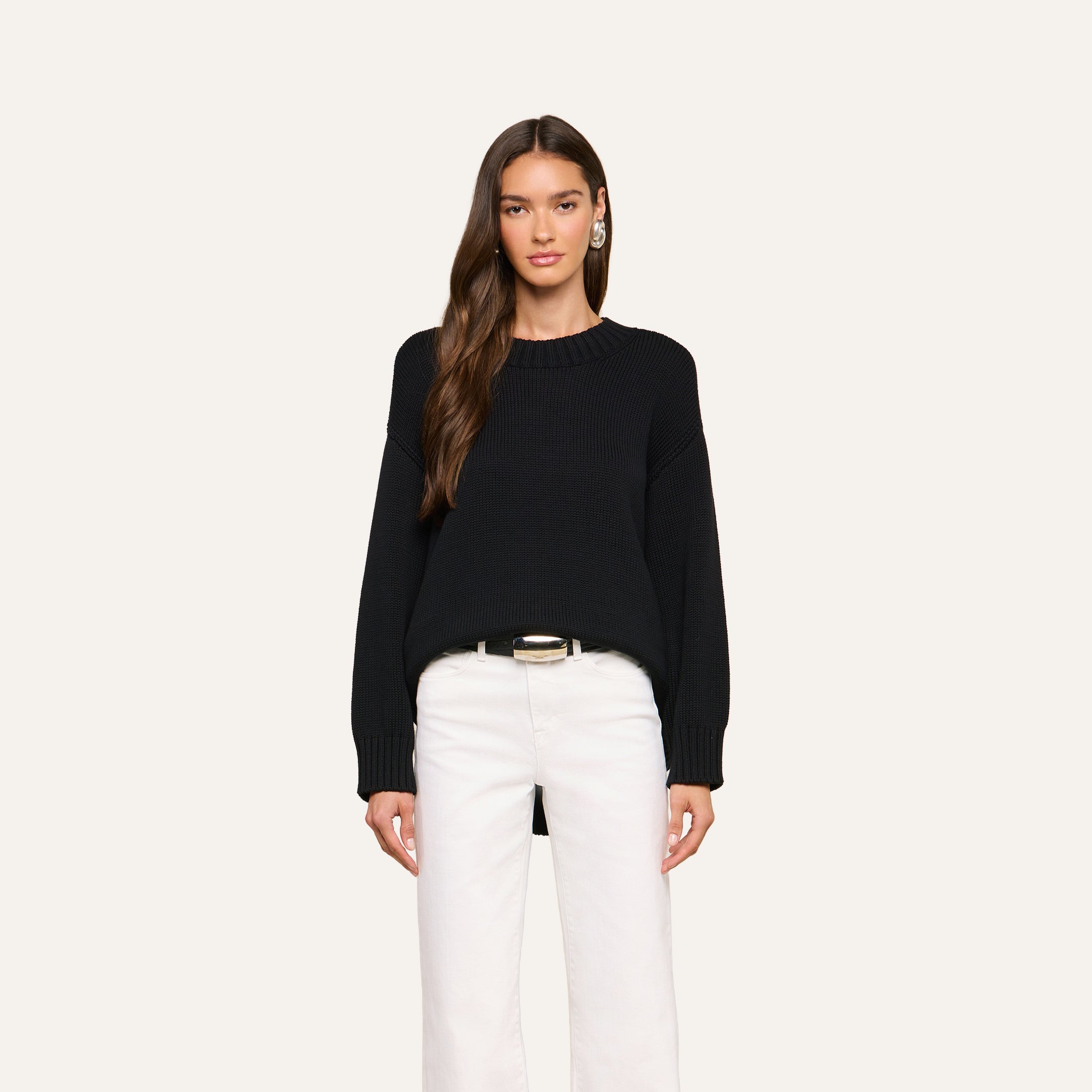 A woman stands facing forward, wearing the Loden Tunic Pullover in black, paired with white pants and a belt. She has long, wavy brown hair and is against a plain, light background.