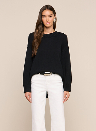A woman stands facing forward, wearing the relaxed fit black Loden Pullover, white pants, and a black belt with a gold buckle. She has long, wavy hair and is set against a plain beige background.