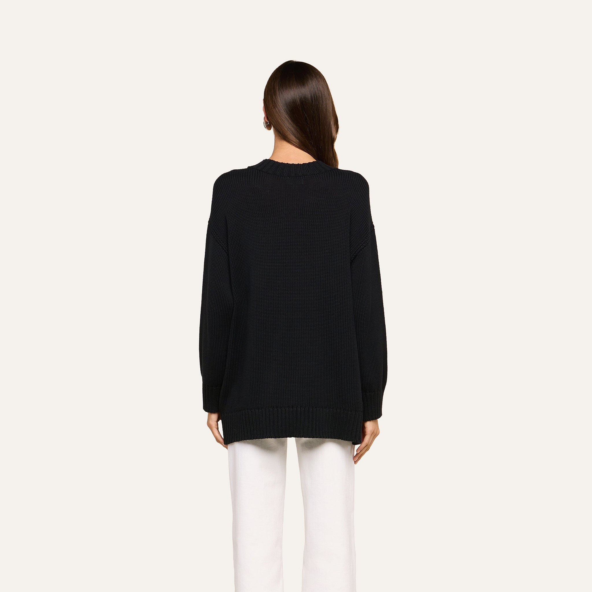 A person with long brown hair, facing away, is wearing a relaxed fit black Loden Tunic Pullover paired with white pants against a plain light background.