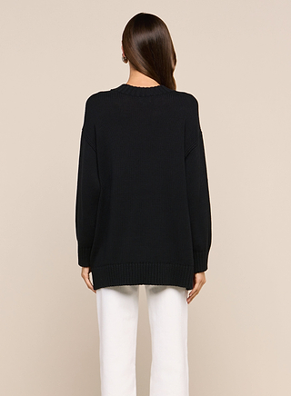 A person with long brown hair wears the Loden Pullover, a relaxed fit black everyday sweater, paired with white pants and standing against a plain beige background, facing away from the camera.