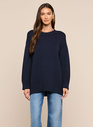 A woman with long brown hair wears the Loden Pullover, a relaxed fit navy blue crewneck tunic sweater, along with light blue jeans, standing against a plain beige background.