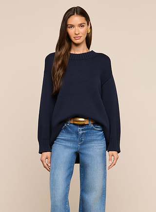 A woman with long brown hair wears the Loden Pullover in navy blue and blue jeans, standing against a plain beige background and facing forward with a relaxed expression.