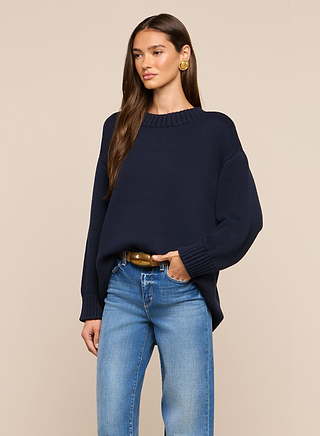 A woman with long brown hair wears the Loden Pullover, styled in a relaxed fit navy shade with blue jeans, a brown belt, and gold earrings, standing against a plain beige background.