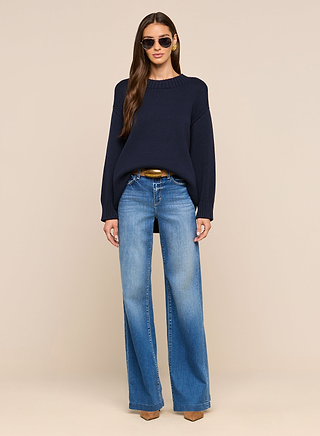 A woman stands against a beige background, facing forward with her hands at her sides, wearing sunglasses and the Loden Pullover—a relaxed fit, dark blue crewneck tunic—paired with light blue wide-leg jeans, a brown belt, and brown pointed shoes.