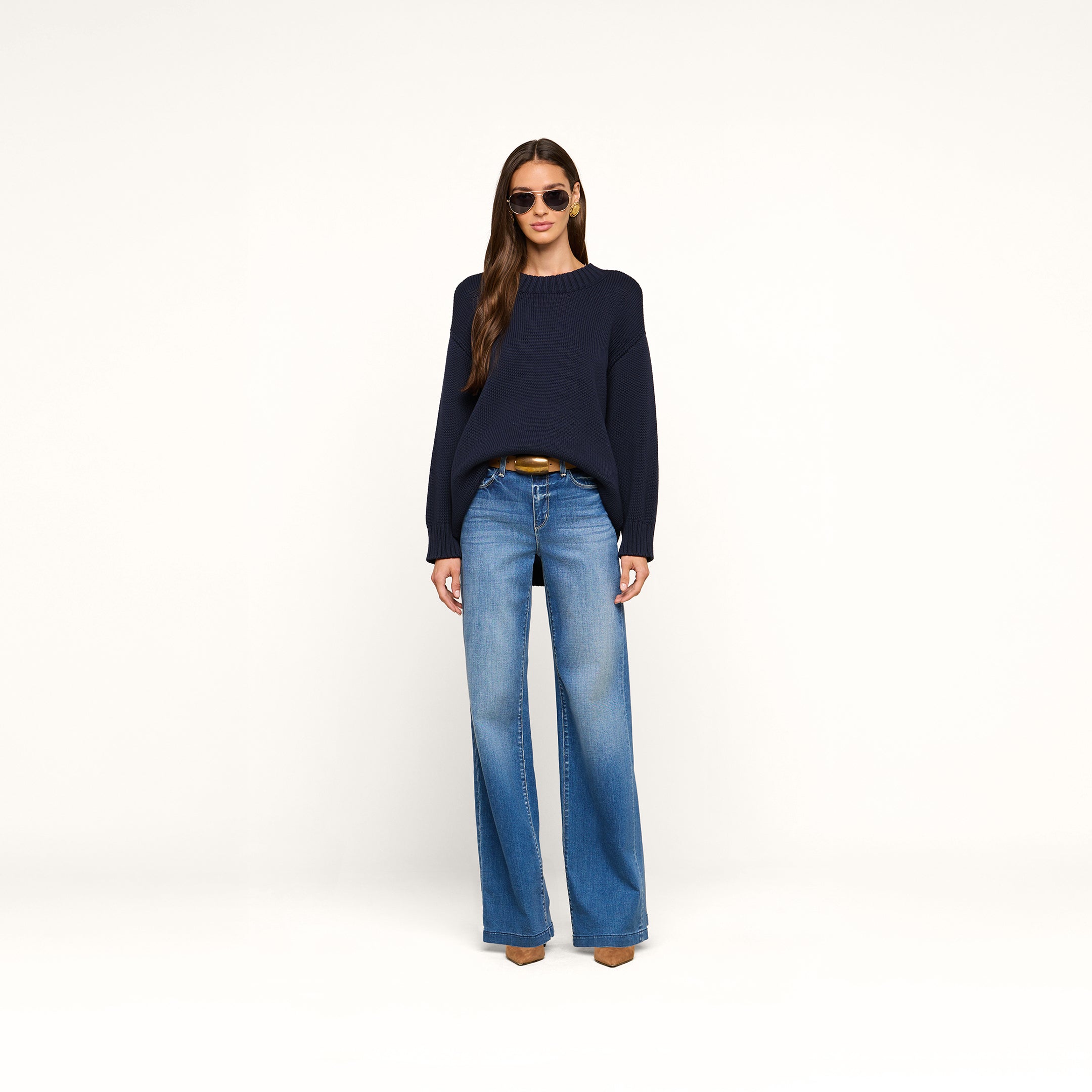 A woman wearing sunglasses and the Loden Tunic Pullover—a navy blue cotton sweater—paired with wide-leg blue jeans, a brown belt, and brown shoes stands against a plain white background, facing forward with her hands by her sides.