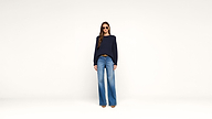 Woman wearing sunglasses, a navy sweater, blue wide-leg jeans, and brown shoes stands against a plain white background.