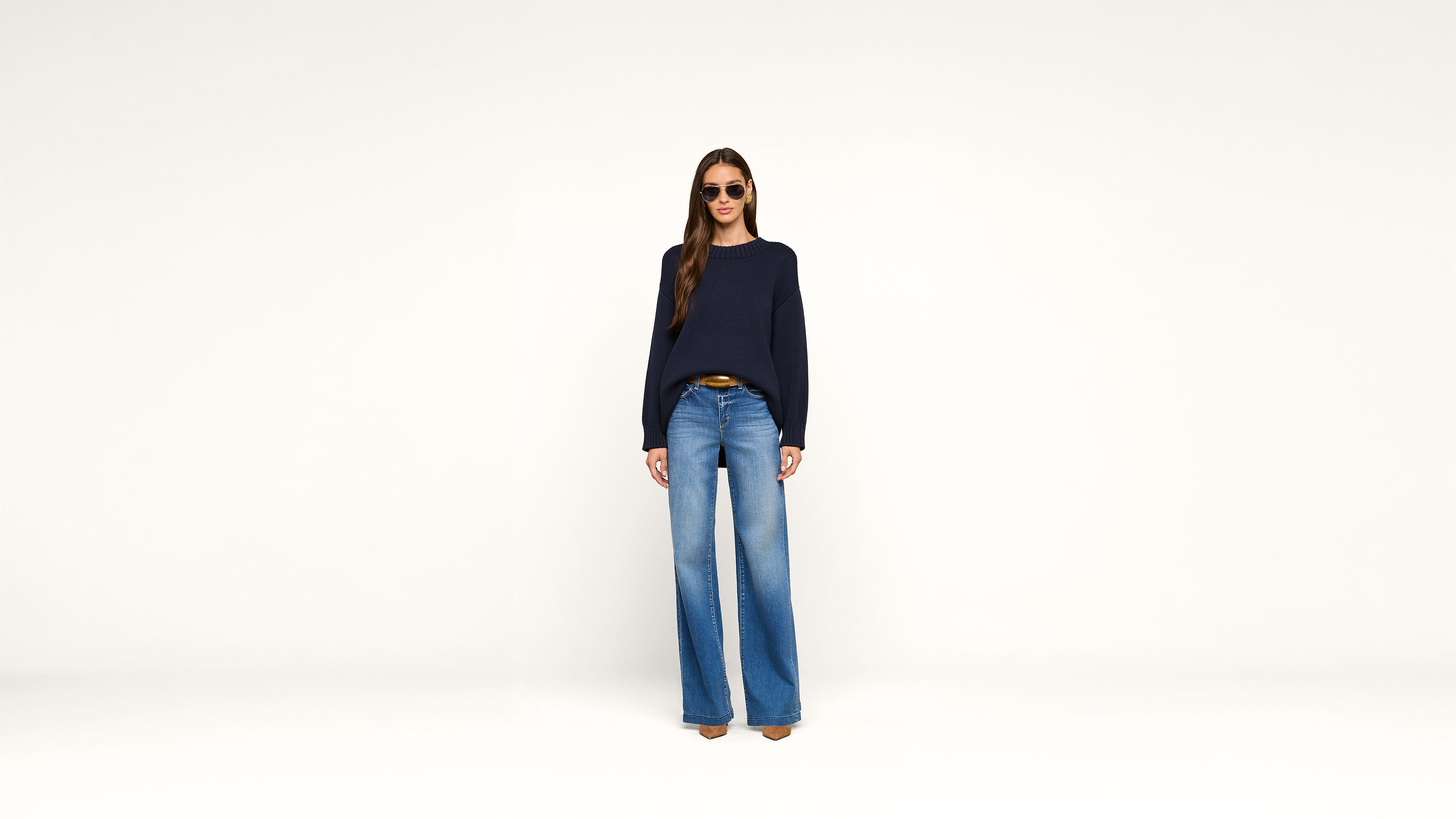 Woman wearing sunglasses, a navy sweater, blue wide-leg jeans, and brown shoes stands against a plain white background.