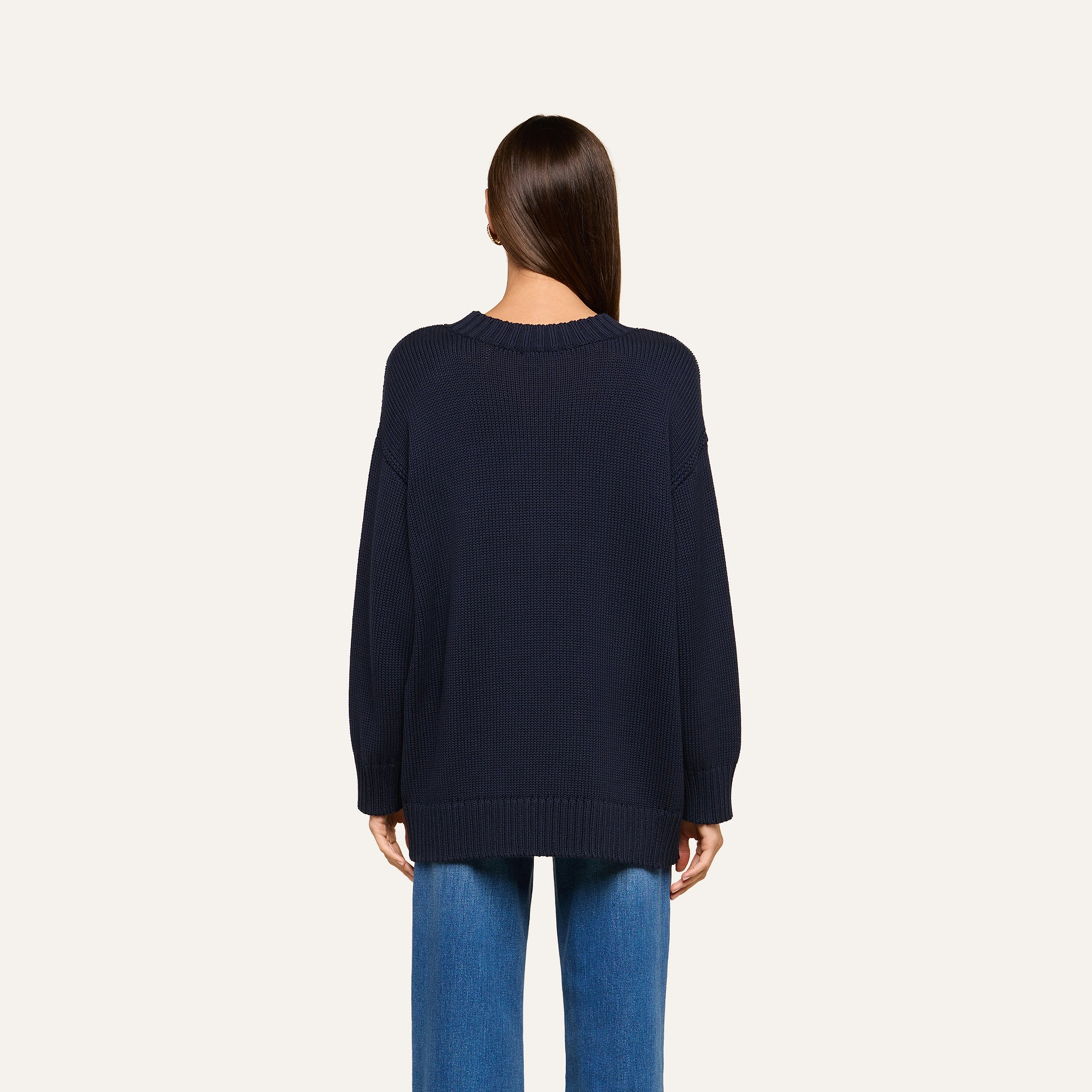 A person with straight brown hair is shown from behind, wearing a loose-fitting navy blue Loden Tunic Pullover and blue jeans against a plain light background.