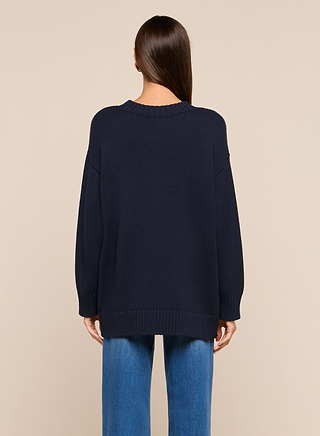 A person with long brown hair is standing with their back to the camera, wearing the Loden Pullover—a relaxed fit, dark navy crewneck tunic sweater—styled with blue jeans against a plain beige background.