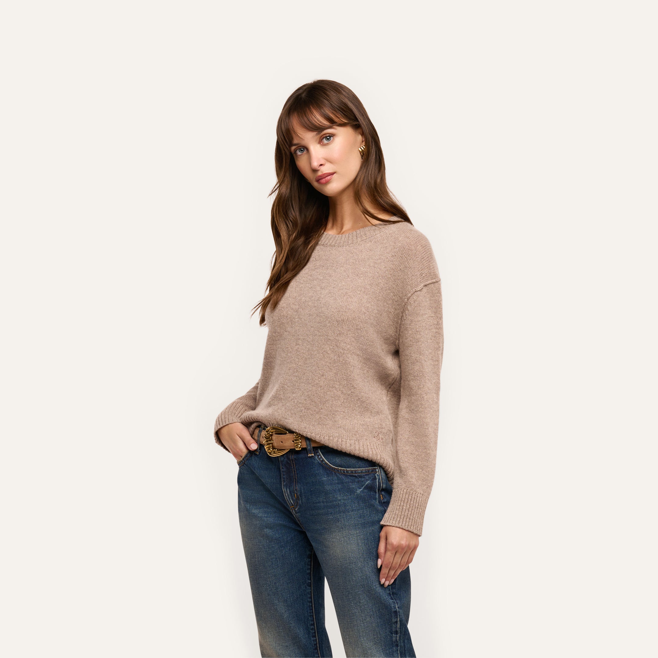 A woman wears a relaxed-fit, beige long-sleeved mid-weight Laine Cashmere Pullover tucked into blue jeans with a decorative belt.