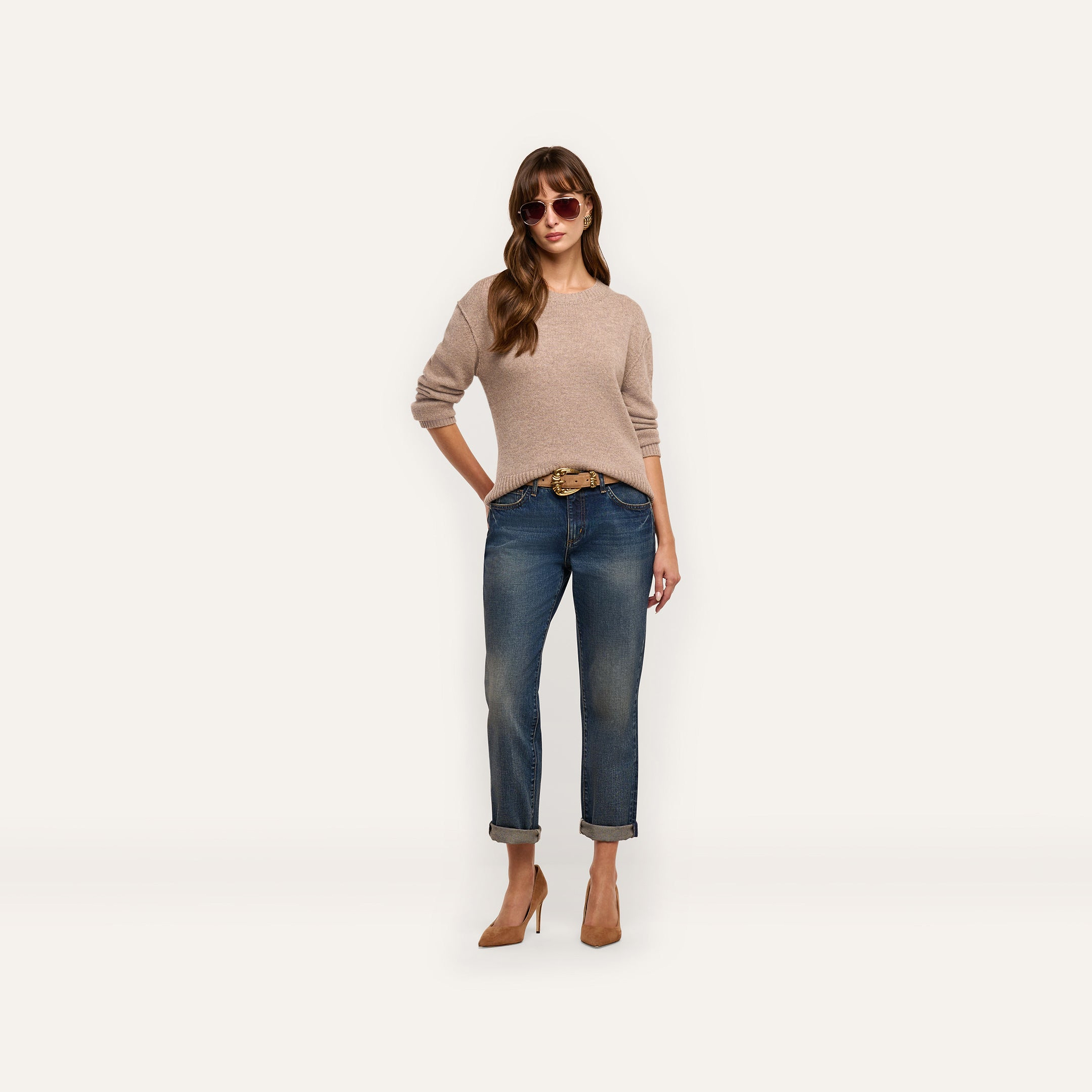 A woman wearing a relaxed fit beige long-sleeved Laine Cashmere Pullover, cuffed blue jeans with a belt, and tan heels, standing against a plain background.