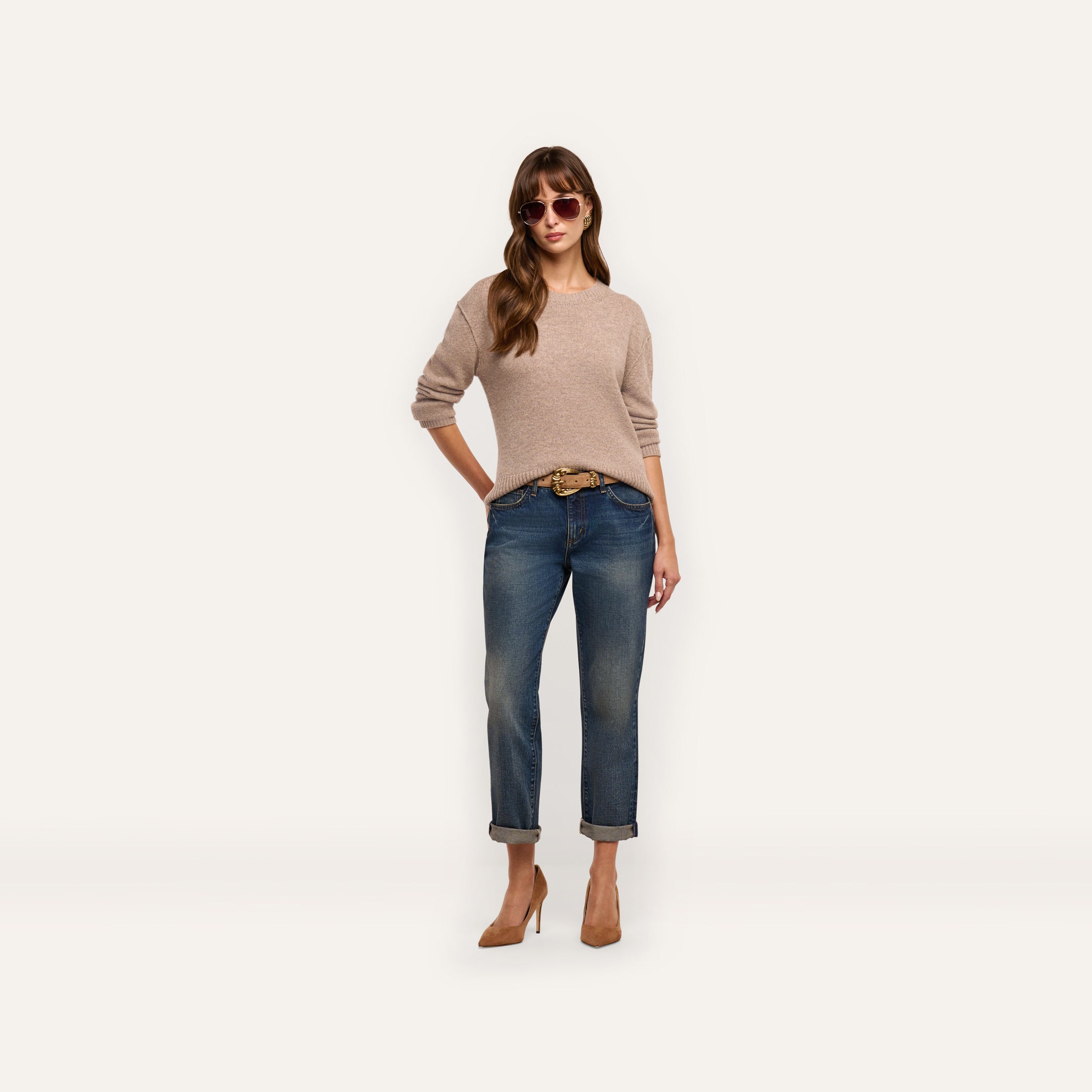 A woman wearing a relaxed fit beige long-sleeved Laine Cashmere Pullover, cuffed blue jeans with a belt, and tan heels, standing against a plain background.