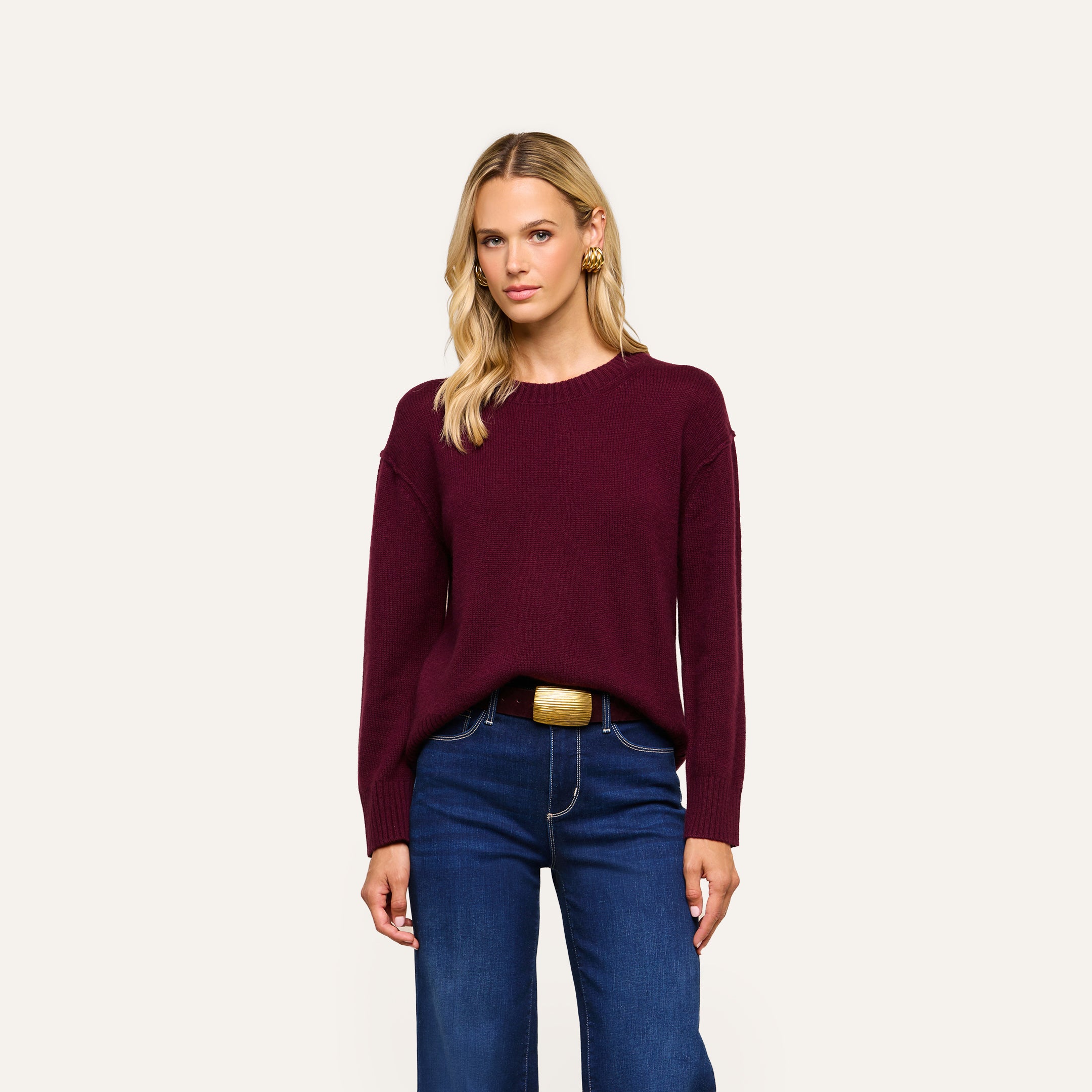 A woman with long blonde hair wearing the Laine Cashmere Top in a relaxed maroon fit, paired with dark blue jeans and a wide belt, stands against a plain light background, looking at the camera with a neutral expression.