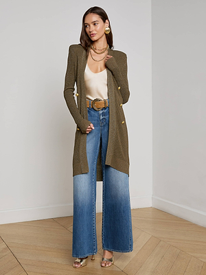A woman stands indoors on a wooden floor wearing the L'AGENCE Noe Double-Breasted Cardigan, a cream satin top, wide-leg blue jeans with a wide tan belt, gold high-heeled sandals, layered necklaces, and large earrings.