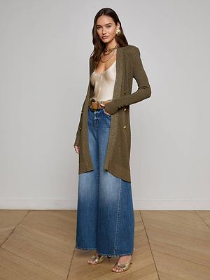 A woman with long wavy hair poses indoors wearing the L'AGENCE Noe Double-Breasted Cardigan, a satin beige camisole, high-waisted wide-leg blue jeans, metallic sandals, and large round earrings.