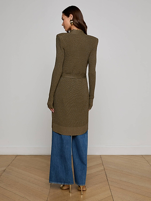 A woman with long brown hair wears the L'AGENCE Noe Double-Breasted Cardigan, styled with wide-leg blue jeans, beige heels, and large earrings. She stands indoors on a wooden floor, facing away from the camera.