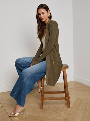 A woman with long brown hair sits on a wooden stool, wearing the L'AGENCE Noe Double-Breasted Cardigan in olive-green over a beige top, blue wide-leg jeans, and gold heels, in a room with light walls and wood flooring.