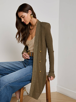 A woman with loose hair looks down while sitting on a wooden stool against a white wall, wearing the L’AGENCE Noe Double-Breasted Cardigan in pine green with subtle sparkle, a tan top, wide-leg blue jeans, gold earrings and heels, and layered necklaces.