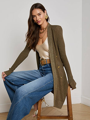 A woman with long brown hair poses on a stool in wide-leg blue jeans, a gold satin top, gold jewelry, statement earrings, beige heels, and the L'AGENCE Noe Double-Breasted Cardigan in pine green before a plain background.