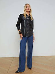 Woman standing against a plain background, wearing the Avina Leather Shirt—a black button-up leather shirt with gold snap buttons—paired with wide-leg blue jeans, black pointed shoes, and bracelets on her left wrist.