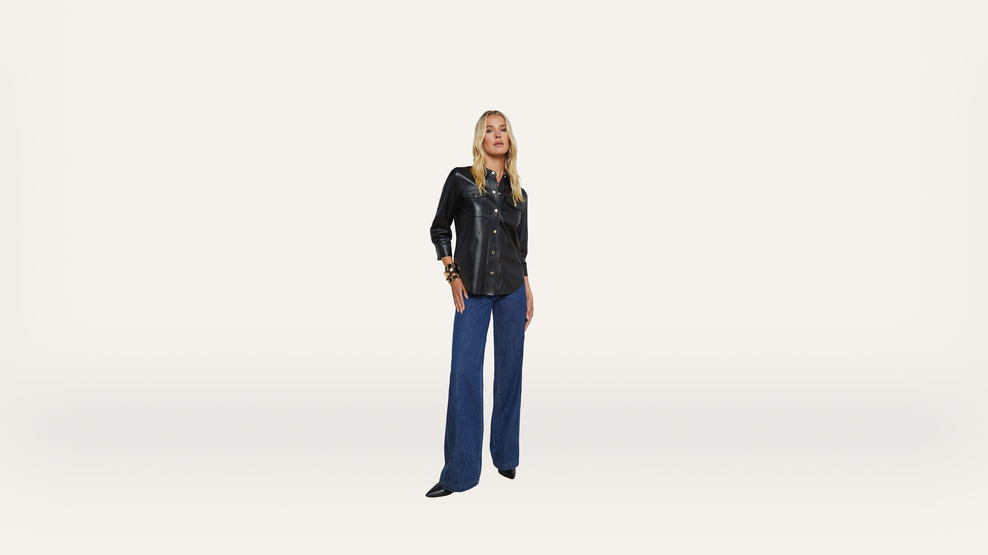 A woman stands against a plain white background, wearing a black long-sleeve shirt and blue wide-leg pants with black shoes.
