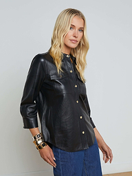 A woman with long blonde hair wears the Avina Leather Shirt, a black lambskin leather button-up featuring chest pockets and gold snap buttons, paired with blue jeans and gold bracelets, standing against a plain light background.