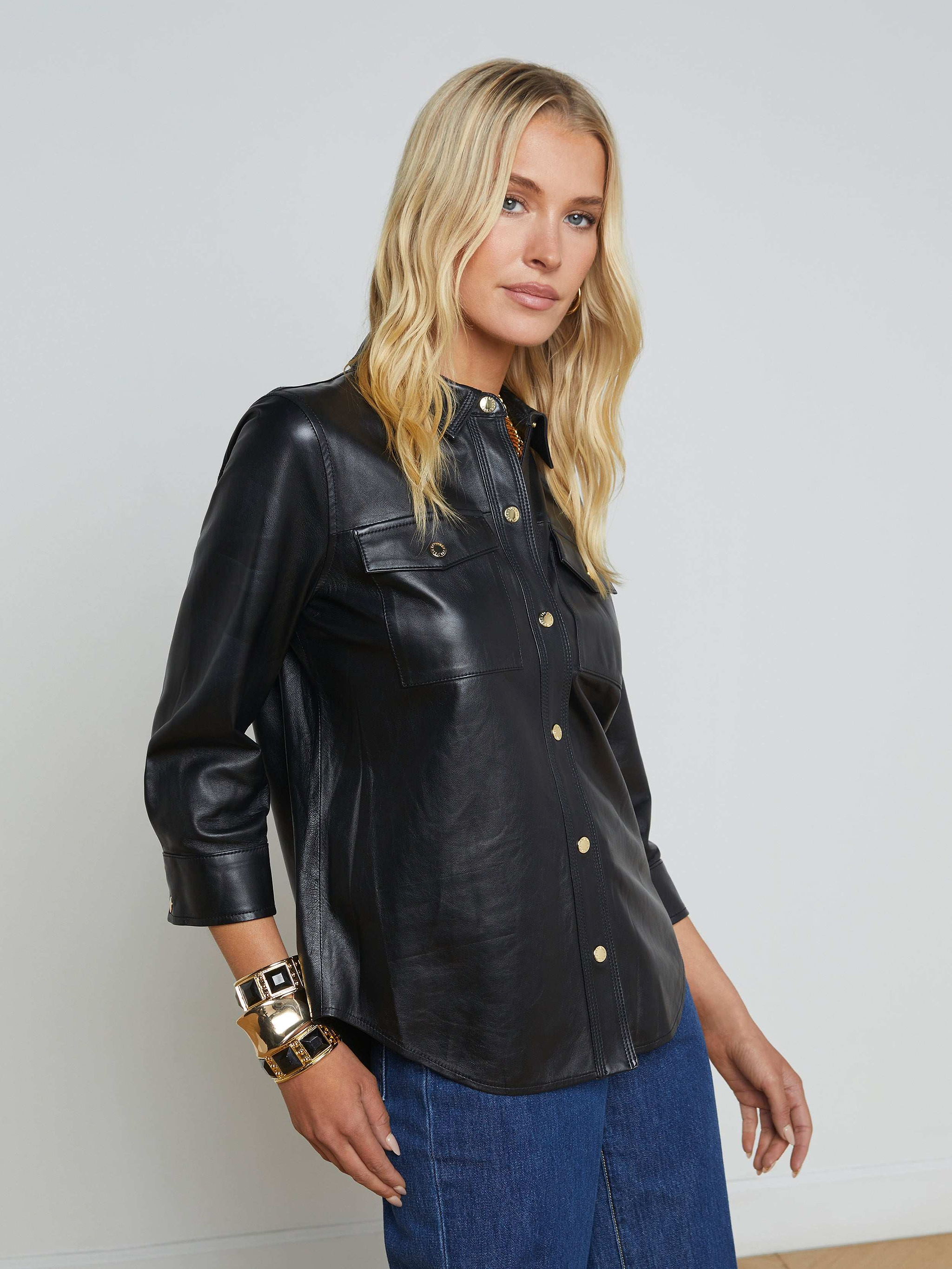 A woman with long blonde hair wears the Avina Leather Shirt, a black lambskin leather button-up featuring chest pockets and gold snap buttons, paired with blue jeans and gold bracelets, standing against a plain light background.