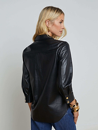 A woman with blonde hair is seen from the back, looking over her shoulder against a plain light background, wearing the Avina Leather Shirt—a black lambskin leather long-sleeve shirt with gold snap buttons—paired with blue jeans and gold bracelets.