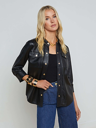 A woman with long blonde hair wears the Avina Leather Shirt—a black lambskin leather button-up with gold snap buttons—layered over a black top, paired with blue jeans and gold jewelry, standing against a plain light background.