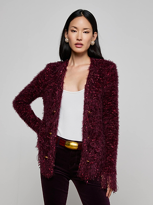 A woman with long black hair wears the L'AGENCE Azure Fringe Cardigan Blazer in deep burgundy with gold buttons over a white top and dark pants, standing confidently with one hand on her hip against a plain, light background.