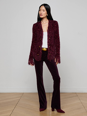 A woman stands against a plain wall in the L'AGENCE Azure Fringe Cardigan Blazer, featuring textured burgundy fabric and gold buttons, paired with matching velvet pants, a white top, burgundy boots, and a gold-buckled belt.