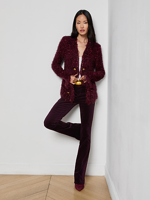 A woman with long black hair wears the L'AGENCE Azure Fringe Cardigan Blazer over a light top and matching burgundy velvet pants with pointed shoes, standing against a white wall on a wooden floor.