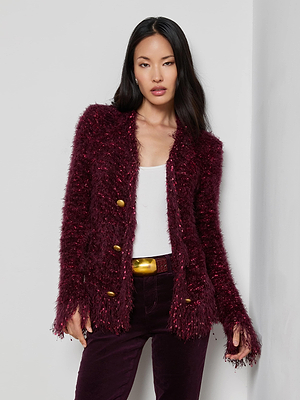 A woman with long dark hair wears the L'AGENCE Azure Fringe Cardigan Blazer in textured burgundy with gold buttons, over a white top and matching burgundy pants, accessorized with a gold belt against a plain white wall.