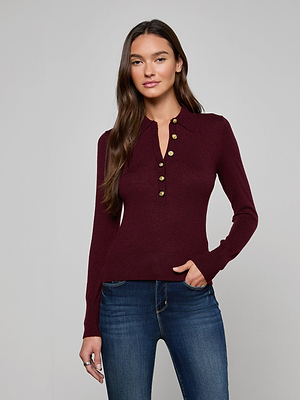 A woman with long brown hair wears the Sterling Sweater, a burgundy premium knit collared henley with gold buttons, paired with blue jeans. She stands against a plain gray background with one hand in her pocket.