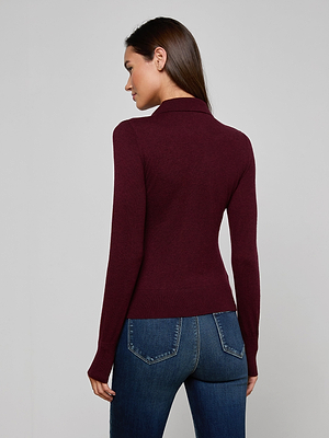 A woman with long brown hair models the Sterling Sweater, a fitted maroon long sleeve knit turtleneck, paired with blue jeans, standing with her back mostly to the camera against a plain gray background.