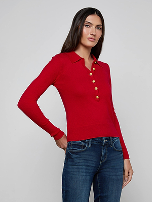 A woman with long dark hair wears the L'AGENCE Sterling Sweater, a slim fit red henley knit top with gold buttons and collar, paired with dark blue jeans, standing against a plain light gray background.