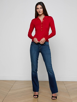 A woman stands indoors on a wooden floor against a white wall, wearing the L'AGENCE Sterling Sweater—a slim fit red knit top with gold buttons and Henley placket—paired with blue jeans and black open-toe heels.
