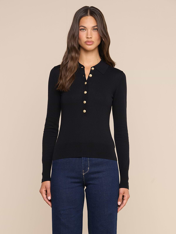 Wearing the Sterling Sweater—a slim fit black Henley top with gold buttons—and dark blue jeans, a woman with long brown hair stands against a plain light background.