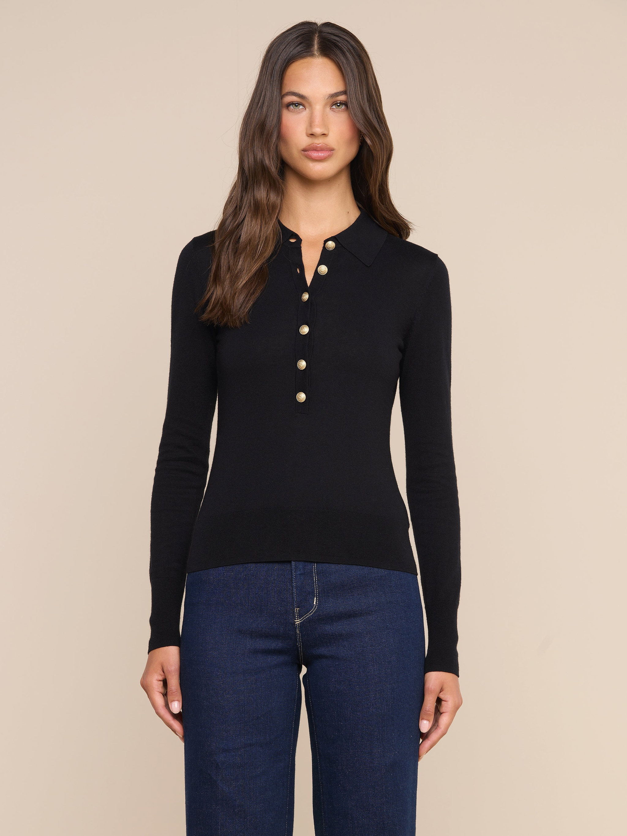 Wearing the Sterling Sweater—a slim fit black Henley top with gold buttons—and dark blue jeans, a woman with long brown hair stands against a plain light background.