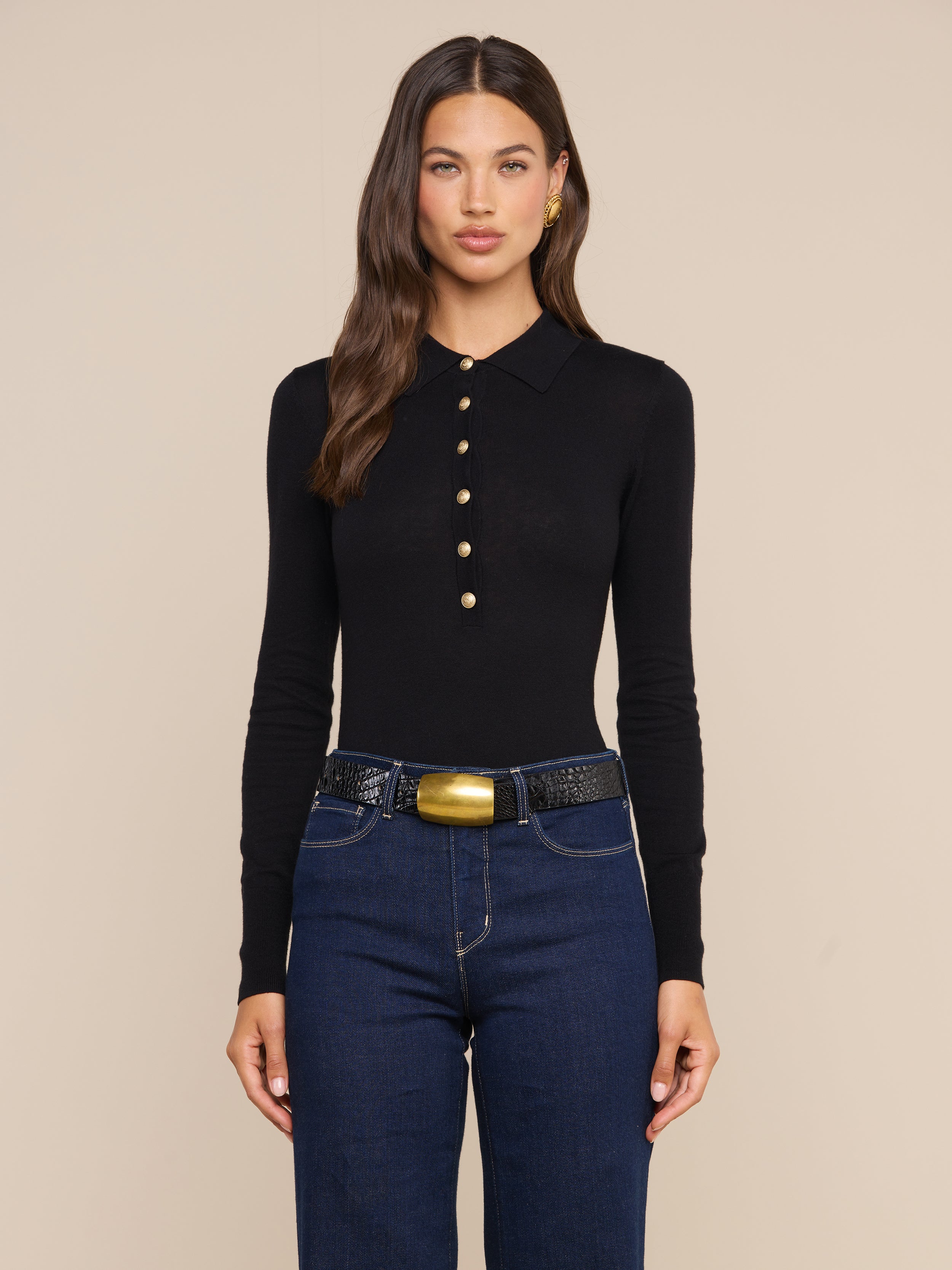 Wearing the Sterling Sweater, a woman with long brown hair pairs it with dark blue jeans and a wide black belt featuring a gold buckle, standing against a plain light background.