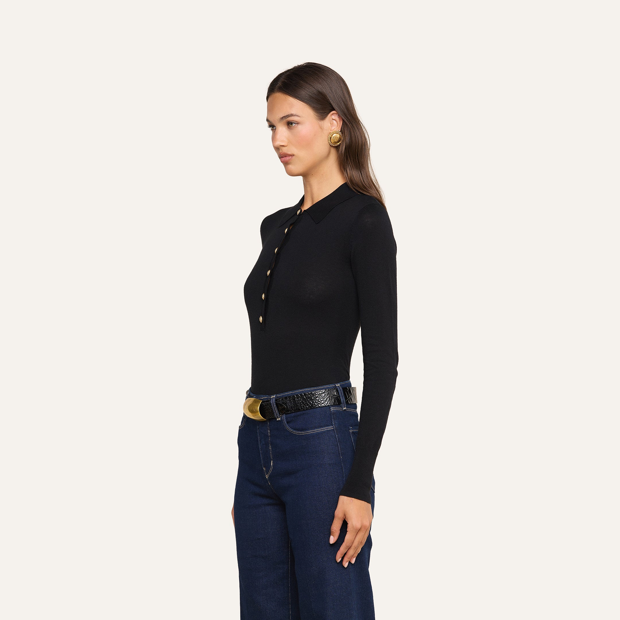 Woman standing in profile, wearing a slim fit black Sterling Sweater with gold buttons, dark blue jeans, a black belt with a gold buckle, and gold earrings, against a plain light background.