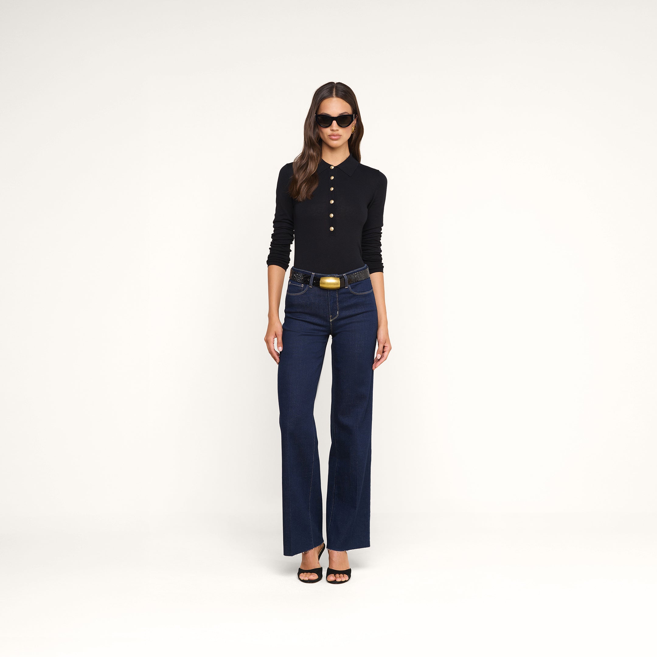 A woman wearing the Sterling Sweater, a black long sleeve knit top, along with dark blue high-waisted jeans, black sandals, sunglasses, and a wide belt stands against a plain white background.